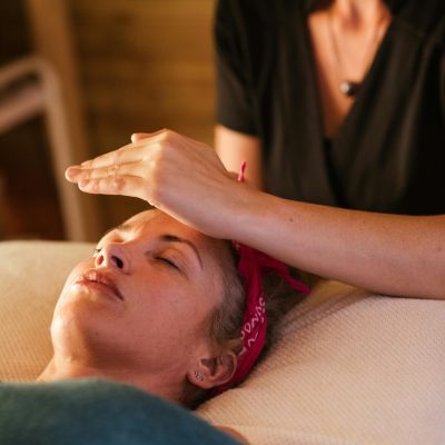 Relaxed woman with closed eyes lying under blanket while getting reiki therapy treatment from unrecognizable therapist in cozy salon with blurred background